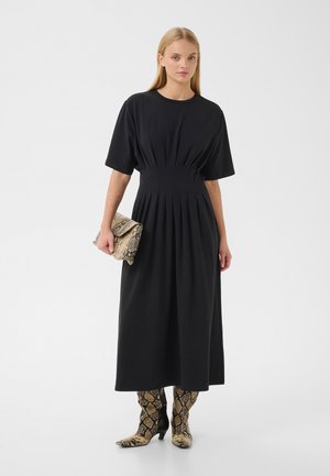 Kvinde iført en sort plisseret midi-kjole med korte ærmer, der holder en clutch med slangeprint og har matchende knælange støvler med slangeprint på.