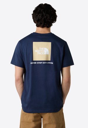 Man draagt een navy blauwe T-shirt met een beige "The North Face" logo en de tekst "Never Stop Exploring", gecombineerd met beige broek, gezien van achteren.