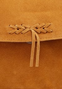 Sac en daim beige au toucher lisse, avec un détail tressé en haut et deux pampilles en daim suspendues. Fermoir circulaire en métal visible.