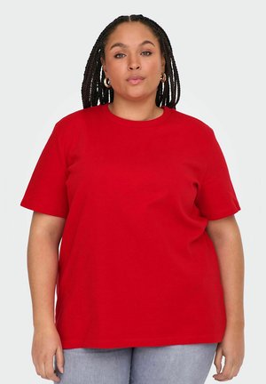 Femme aux cheveux tressés portant des boucles d'oreilles en anneau, un t-shirt rouge à manches courtes et un pantalon gris clair, debout devant un fond uni.