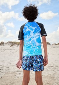 Sort badedragtstop med blåt vandplaskedesign, kombineret med blå mønstrede shorts. Struktureret stof, afslappet sportsligt fit.