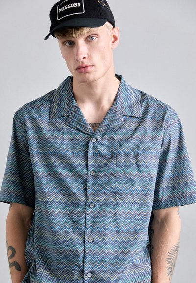 Blonds vīrietis valkā zīmulēku rakstu zilu krekla ar pogām un melnu cepuri ar "MISSONI" logotipu, parādot tetovējumus uz rokām un krūtīm.