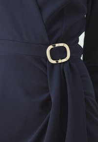 Námorná oblečená sukňa so štruktúrovanou látkou, s zlatou obdĺžnikovou prackou v páse a drapériou na jednej strane.