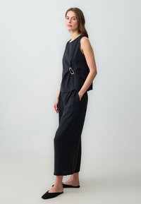 Tenue noire comprenant un haut sans manches avec un twist sur le côté et un grand détail en anneau métallique, associé à un pantalon cropped ample et des chaussures à enfiler.