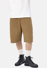 Cuerpo inferior de una persona que lleva pantalones cortos marrones hasta la rodilla, calcetines blancos de caña corta, botas de combate negras con cordones y una camiseta blanca.