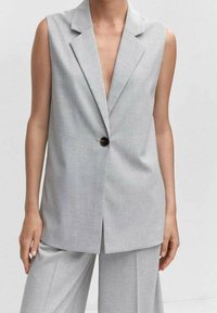 Bezrukávý světle šedý tailoringový blejzr s jedným černým knoflíkem, nosí se s odpovídajícími vysokými širokými kalhotami.