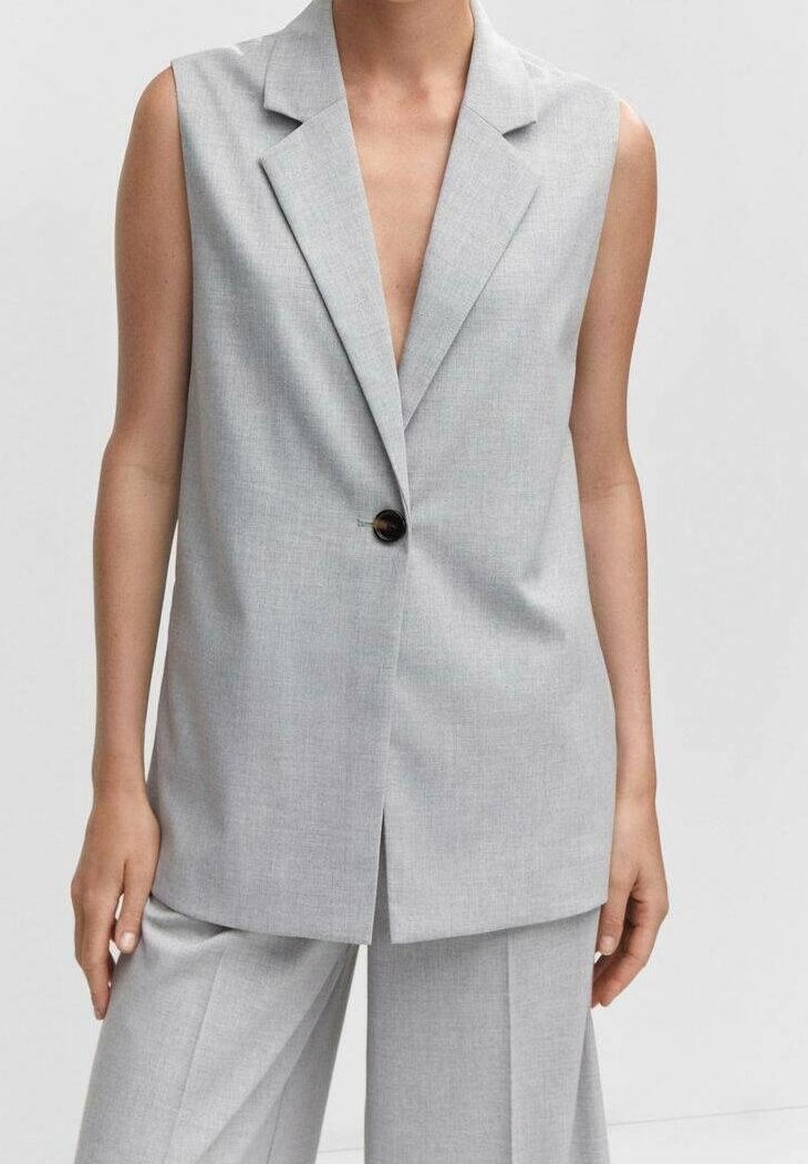 Bezrukávý světle šedý tailoringový blejzr s jedným černým knoflíkem, nosí se s odpovídajícími vysokými širokými kalhotami.