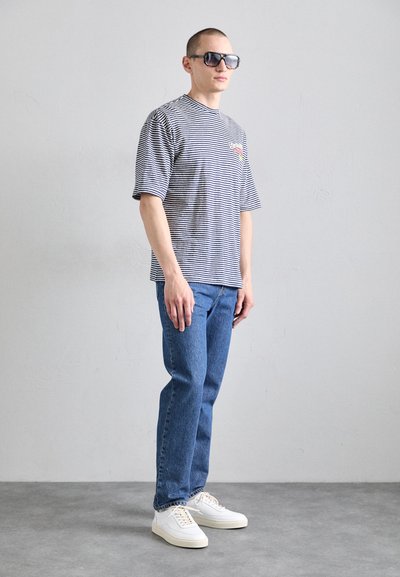 Gestreiftes kurzärmliges T-Shirt in Schwarz und Weiß, blaue Jeans und weiße Sneakers. Zudem mit übergroßen schwarzen Sonnenbrillen und einem bunten Logo ausgestattet.