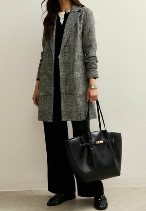 Femme portant un manteau à carreaux gris par-dessus une tenue noire, tenant un grand sac fourre-tout en cuir noir, avec une montre-bracelet dorée visible au poignet gauche.