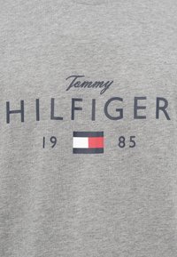 Sivi materijal s natpisom "Tommy Hilfiger 1985" i pravokutnim logom sa bijelim, crvenim i tamnoplavim blokovima centriranim ispod natpisa.