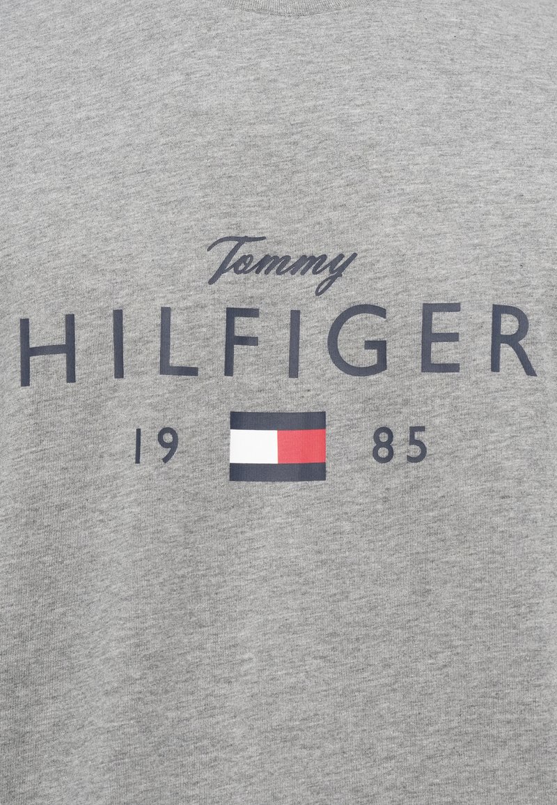 Grå stof med teksten "Tommy Hilfiger 1985" og et rektangulært logo med hvide, røde og marineblå blokke centreret under teksten.