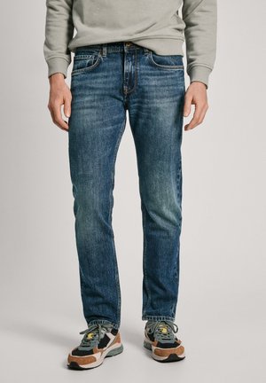 Homme portant un jean bleu, un sweatshirt gris et des baskets multicolores, debout avec les mains relâchées le long du corps devant un fond uni.