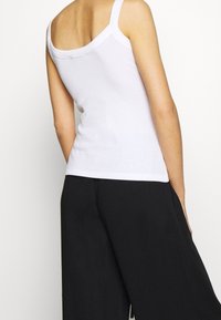 Bílý top bez rukávů s výstřihem do oblouku a žebrovanou strukturou, kombinovaný s černými kalhotami s širokými nohavicemi. Pohled zezadu ukazuje ramenní popruhy.