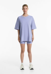 Světležluté oversized tričko s krátkým rukávem, spárované s odpovídajícími sportovními šortkami. Nosené s bílými sportovními botami, na bílém pozadí.