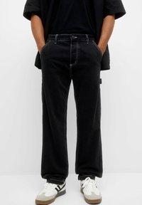 Persona vistiendo pantalones sueltos negros con costuras blancas, manos en los bolsillos, combinados con zapatillas blancas con rayas negras sobre un fondo blanco.