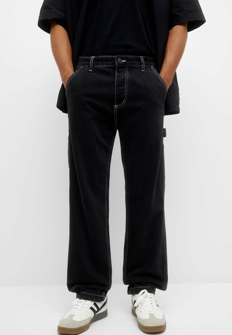 Persona vistiendo pantalones sueltos negros con costuras blancas, manos en los bolsillos, combinados con zapatillas blancas con rayas negras sobre un fondo blanco.