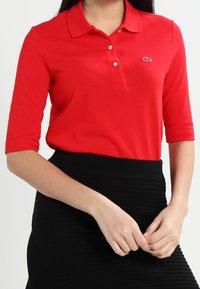 Žena v červeném polo tričku s krátkým rukávem a malým logem krokodýla, s černou žebrovanou sukní, se založenýma rukama před sebou.