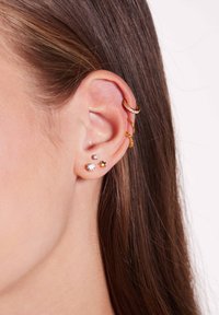 Goudkleurige oorbellen getoond aan het oor; omvat een ring met een kristalaccent, twee studs en een stervormig ontwerp. Glad oppervlak en glans.