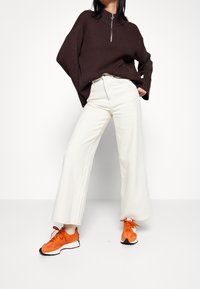 Braun gerippter Pullover mit hohem Kragen und Reißverschluss, kombiniert mit weißen, weiten Hosen und leuchtend orangefarbenen Turnschuhen. Strukturiertes Material.