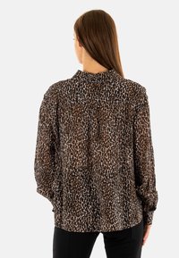Blusa de estampado de leopardo en tejido ligero, con cuello tipo polo, mangas largas y detalle de pliegues en la parte trasera. Los tonos marrón y beige predominan.