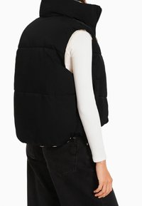 Black puffer vest with high collar, quilted design, and side pockets, worn over a white long-sleeve top, featuring a drawstring hem.