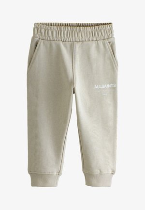 Bambino che indossa pantaloni della tuta beige AllSaints, sneakers bianche e t-shirt bianca, in piedi davanti a uno sfondo grigio uniforme.