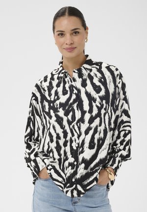 Femeie purtând o cămașă cu nasturi, cu imprimeu de zebru alb-negru, blugi albastru deschis, cu mâinile în buzunare și cercei și brățară aurii hoop.