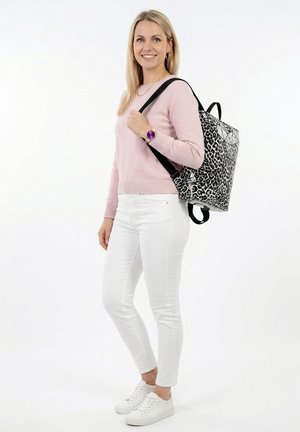 Lächelnde Frau mit einem rosa Pullover, weißen Hosen, weißen Turnschuhen und einem schwarz-weißen Rucksack mit Muster auf der Schulter.