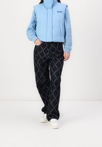 Helles blaues Steppwesten mit hohem Kragen, kombiniert mit schwarz gemusterten Hosen mit Diamantdesign und weißen Sneakers.