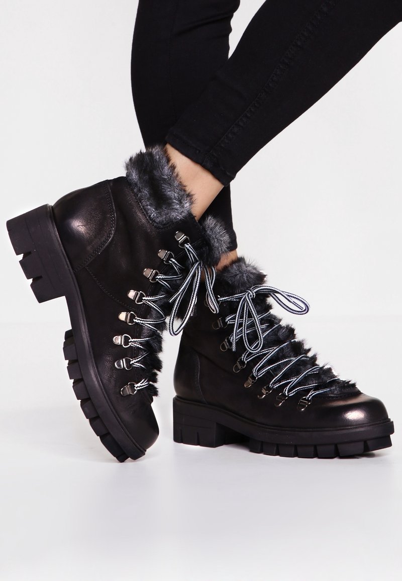 Bottes cheville en cuir noir avec semelles épaisses, doublure en fourrure grise, œillets en métal, et lacets rayés noir et blanc portées avec un pantalon noir.