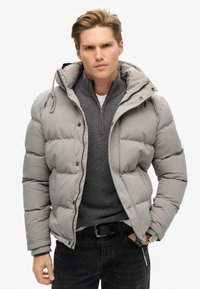 Chaqueta acolchada de color gris claro con capucha, botones de presión y cierre frontal. Llevada sobre un suéter gris oscuro y pantalones negros.