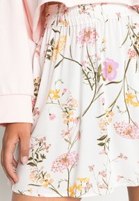Jupe fleurie à base blanche, ornée de fleurs aux couleurs pastel, taille froncée et texture lisse. Main posée sur l'ourlet de la jupe.