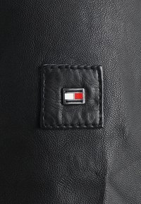 Zwart gestructureerd leer met een gestikte vierkante patch met een klein rechthoekig embleem verdeeld in witte en rode secties.