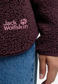 Veste en polaire de couleur bordeaux foncé avec un extérieur texturé et velouté. Présente une broderie rose "Jack Wolfskin" et un logo d'empreinte de patte.