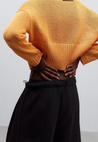 Roter, kurzer Strickpullover mit strukturiertem Muster und weiten Ärmeln, kombiniert mit schwarzen, hochgeschnittenen Hosen mit Kordelzug.