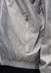 Ανοιχτό γκρι μπουφάν με επαναλαμβανόμενο μοτίβο "AX", κάθετη τσέπη με φερμουάρ και ελαστική μέση, εμφανίζεται στο σώμα.