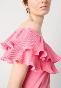 Vestido rosa con escote off-the-shoulder que presenta grandes volantes plisados ondulados. Textura de tela suave con una sección del cuerpo ajustada. Detalle de pendientes de aro plateados.