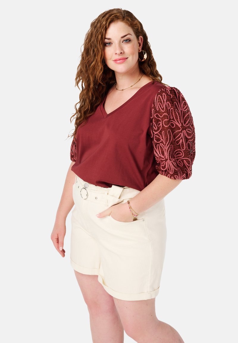 Haut V en bordeaux avec des manches bouffantes rose brodées, associé à un short blanc à revers. Comprend une ceinture et des poches latérales.