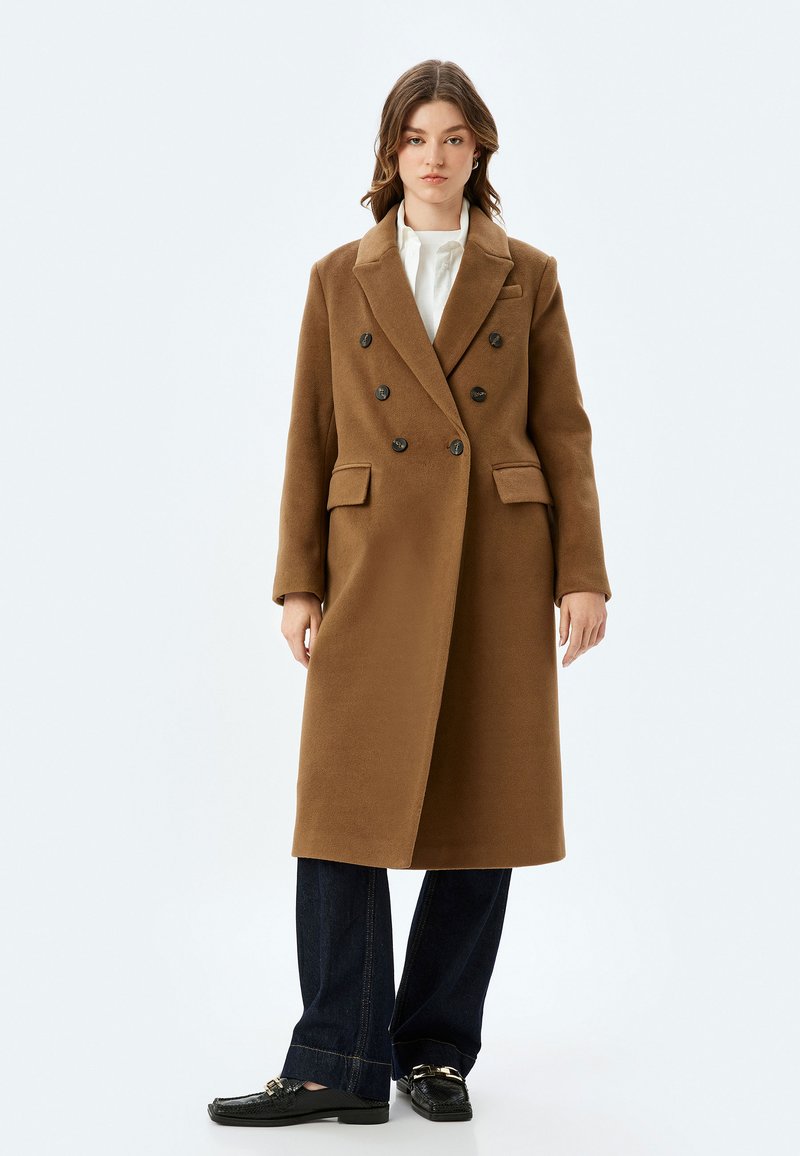Manteau double boutonnage en mélange de laine marron avec de grands boutons noirs, revers crantés, deux poches avant et une coupe ajustée.