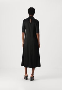Rochie midi neagră, cu mâneci scurte, cu o tăietură în formă de cheie la spate și o siluetă fluidă, fabricată dintr-un material neted. Purtați cu pantofi cu toc pătrat.