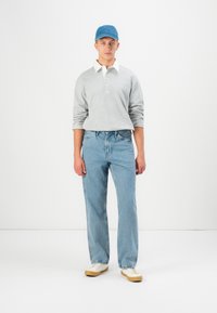 Pelēks garām piedurknēm polo krekls ar baltu apkakli, gaiši zili džinsi un zila cepure. Modelis valkā bēšas sporta kurpes ar baltām kurpju auklām uz viendabīga fona.