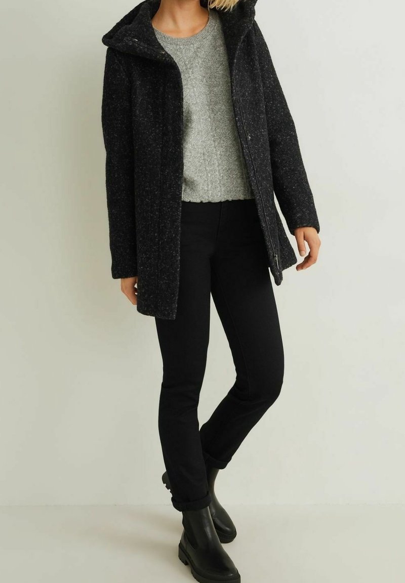 Manteau en mélange de laine noire avec capuche, pull gris en maille, jean noir ajusté et bottines noires. Le manteau est doté d'une fermeture éclair et d'un tissu texturé.