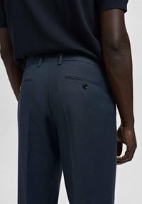 Hombre vestido con pantalones oscuros azul marino y camisa a juego, mostrado desde la cintura hasta la mitad del muslo, de pie contra un fondo claro y liso.