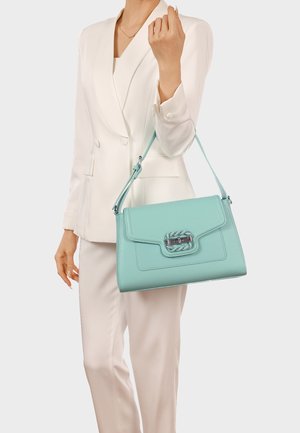 Donna in completo bianco che tiene una borsa a tracolla in pelle azzurro chiaro con fibbia d'argento, su sfondo chiaro e uniforme.
