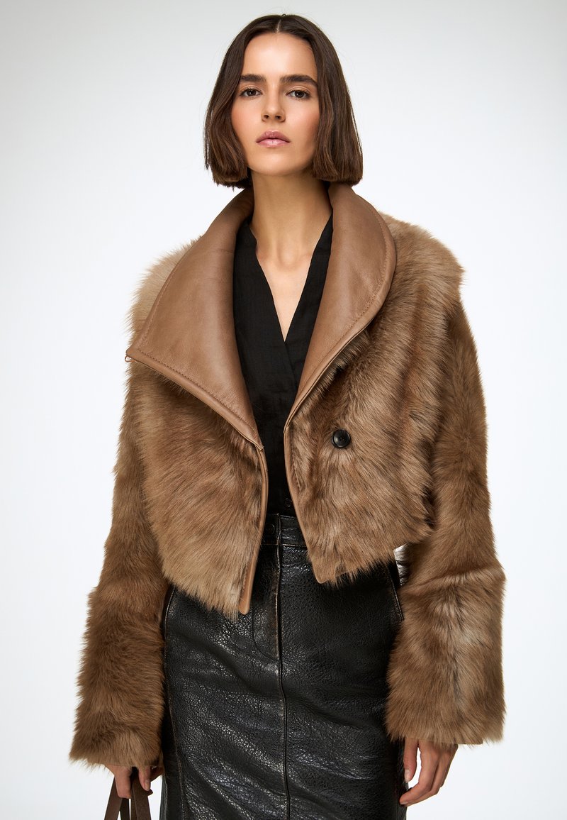 Veste courte en fausse fourrure marron douce avec un col en cuir, dotée d'une fermeture à un seul bouton et de manches larges, portée sur un haut noir.