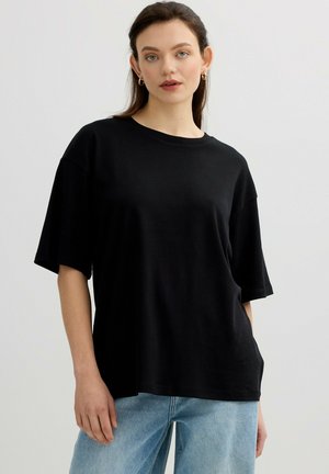 Joven mujer con cabello largo y castaño que lleva pendientes de aro dorados, una camiseta negra de manga corta y suelta, y vaqueros azul claro, de pie frente a un fondo liso.