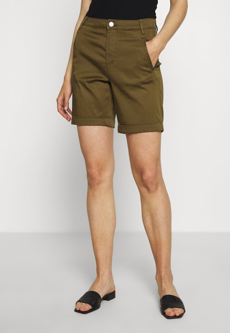 Personne portant un short vert olive longueur genou avec ourlets retournés, un haut noir sans manches et des chaussures noires ouvertes à l'avant, debout avec une main dans la poche.