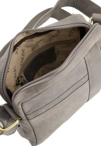 Grå læder crossbody taske med lynlåslukning, indre lommer og et stoffoer med et gentagende logomønster.