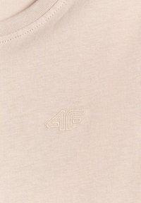 Gros plan d'un tissu beige avec un petit logo brodé "4F" près de l'encolure d'un vêtement à col rond.
