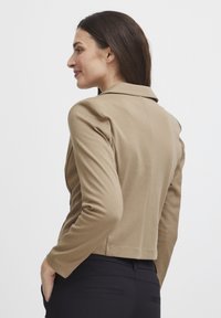 Kvinna med långt brunt hår som bär en beige kavaj och svarta byxor, står på sidan med en hand i fickan mot en vit bakgrund.
